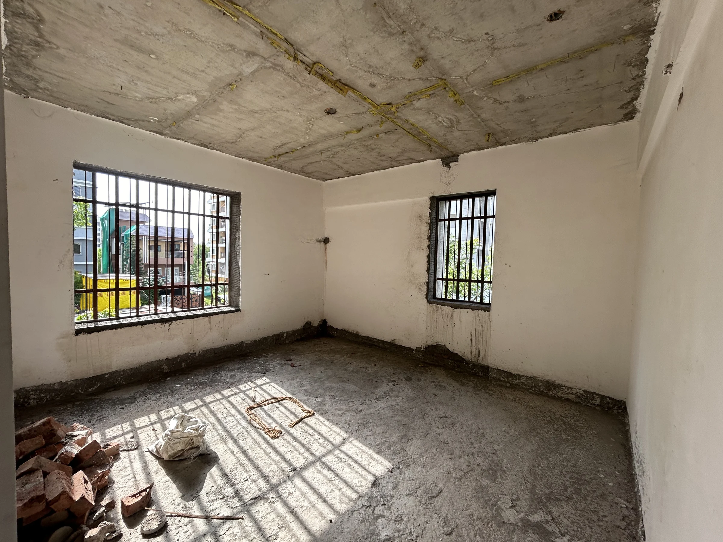 Site photograph: an empty plastered room with two grilled windows and sun on the floor